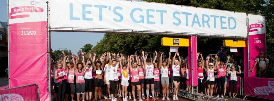 a group of enthusiastic participants raises their hands in celebration under a "LET'S GET STARTED" banner at the Race for Life event