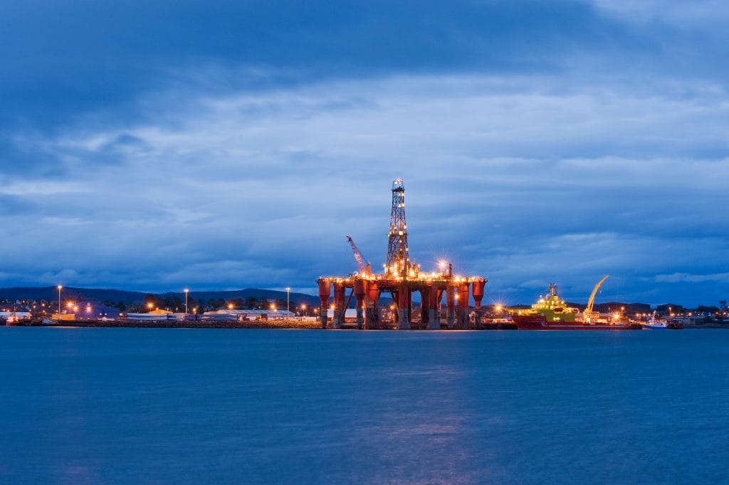 an illuminated offshore oil rig stands in the ocean at dusk, with a nearby vessel and clouds creating a dramatic backdrop