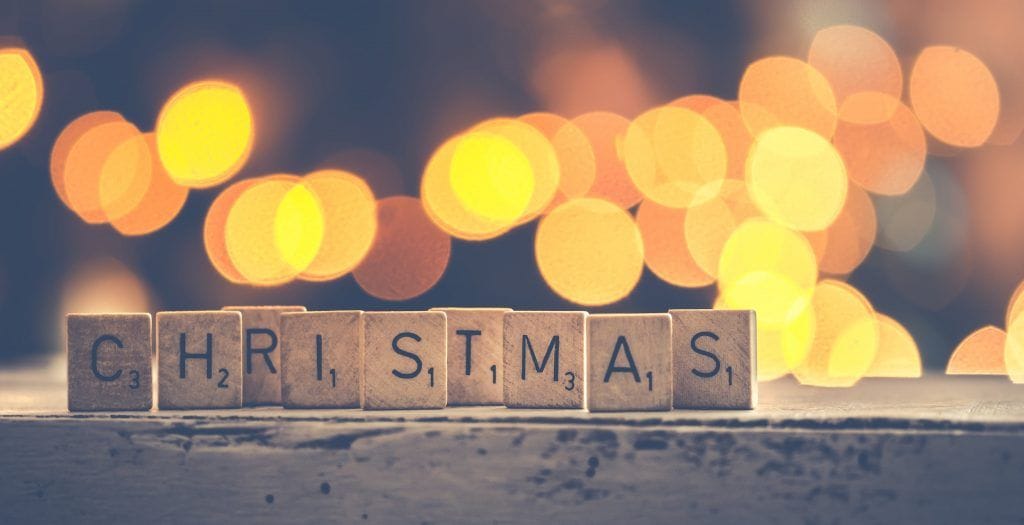 scrabble tiles spell "CHRISTMAS" against a softly blurred backdrop of warm, glowing holiday lights