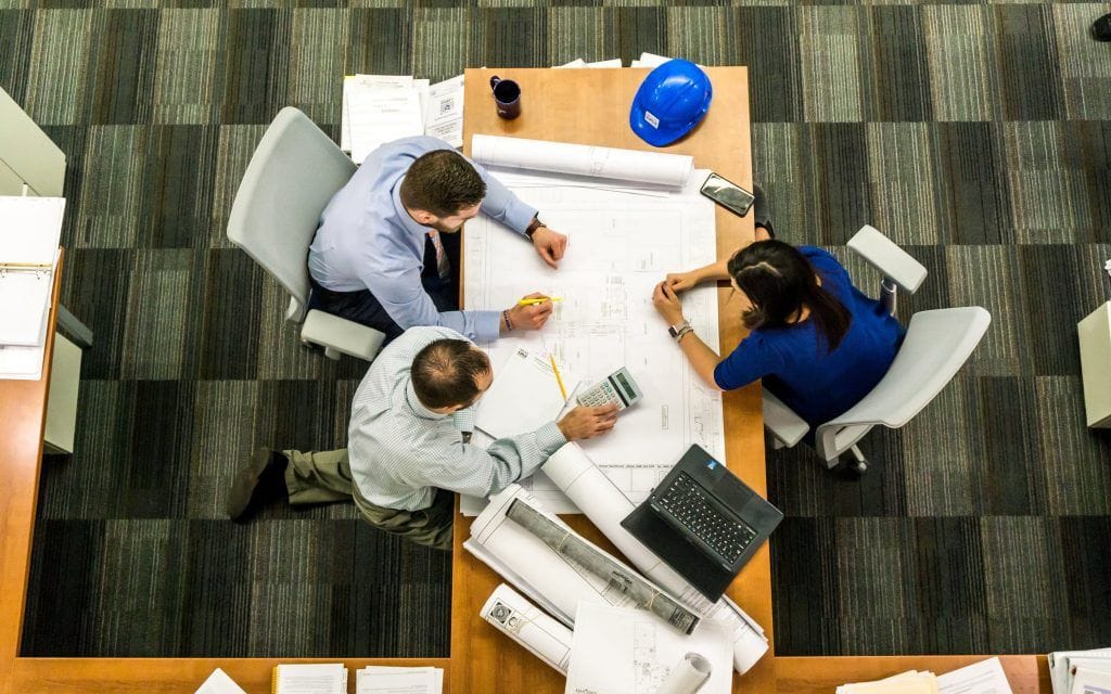 three professionals collaborate over blueprints and plans, using a calculator and laptop in a modern office setting