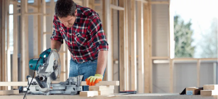 a person in a plaid shirt and gloves operates a power saw, measuring lumber in a wooden frame construction area