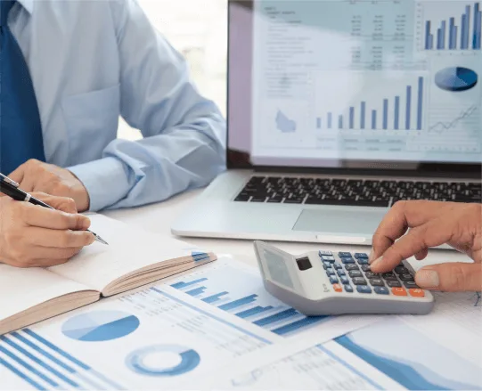 two professionals analyzing financial data, one using a calculator, while a laptop displays charts and graphs on a desk with papers