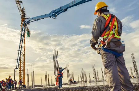 construction workers in safety gear operate machinery at a building site, with rebar columns rising against a sunset sky