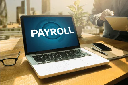 a laptop on a desk displaying "PAYROLL" on the screen, with a plant and a person holding a tablet in the background