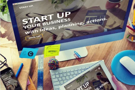 a computer and tablet displaying a business startup website, surrounded by stationery and office supplies on a wooden desk