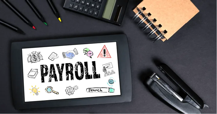 a tablet displaying the word "PAYROLL" surrounded by related icons, on a desk with a calculator, notepad, pens, and a stapler