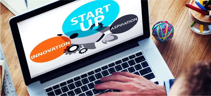 a person's hand on a laptop keyboard displaying "START UP" with keywords "INNOVATION" and "ASPIRATION" on the screen
