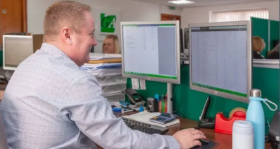 a person in a light blue shirt works at a desk with two monitors, surrounded by office supplies and a water bottle