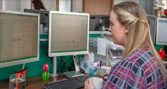 a woman with long, wavy hair sits at a desk, holding a cup and working on dual computer monitors surrounded by office supplies