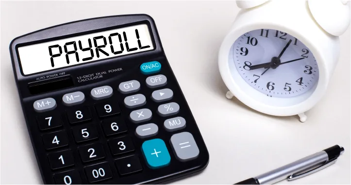 a black calculator displaying "PAYROLL" beside a white alarm clock and a silver pen on a light surface