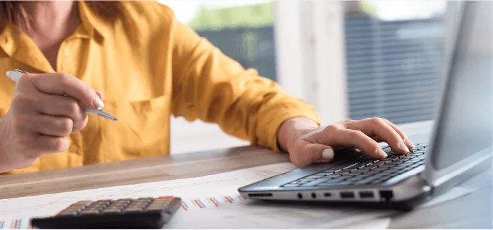 a person in a yellow shirt uses a laptop, holding a pen in one hand while working among financial papers and a calculator