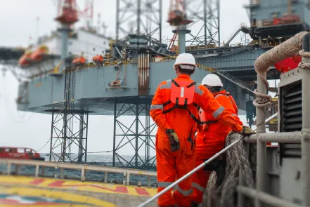 two workers in orange coveralls and helmets stand on a vessel, overlooking an offshore oil rig in a cloudy atmosphere